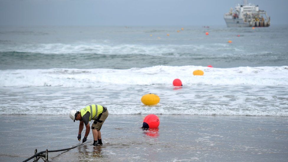 Un trabajador al borde del mar con un barco en alta mar está trabajando en el amarre de un cable de fibra óptica que se ve saliendo del mar, que se extiende entre los EE. UU. y España en la imagen tomada en 2017
